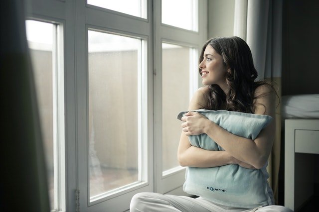a girl sitting and watching out of the window while holding a pillow 